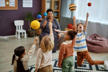 Diverse group of little kids playing with planet models while enjoying class in child care with teacher watching